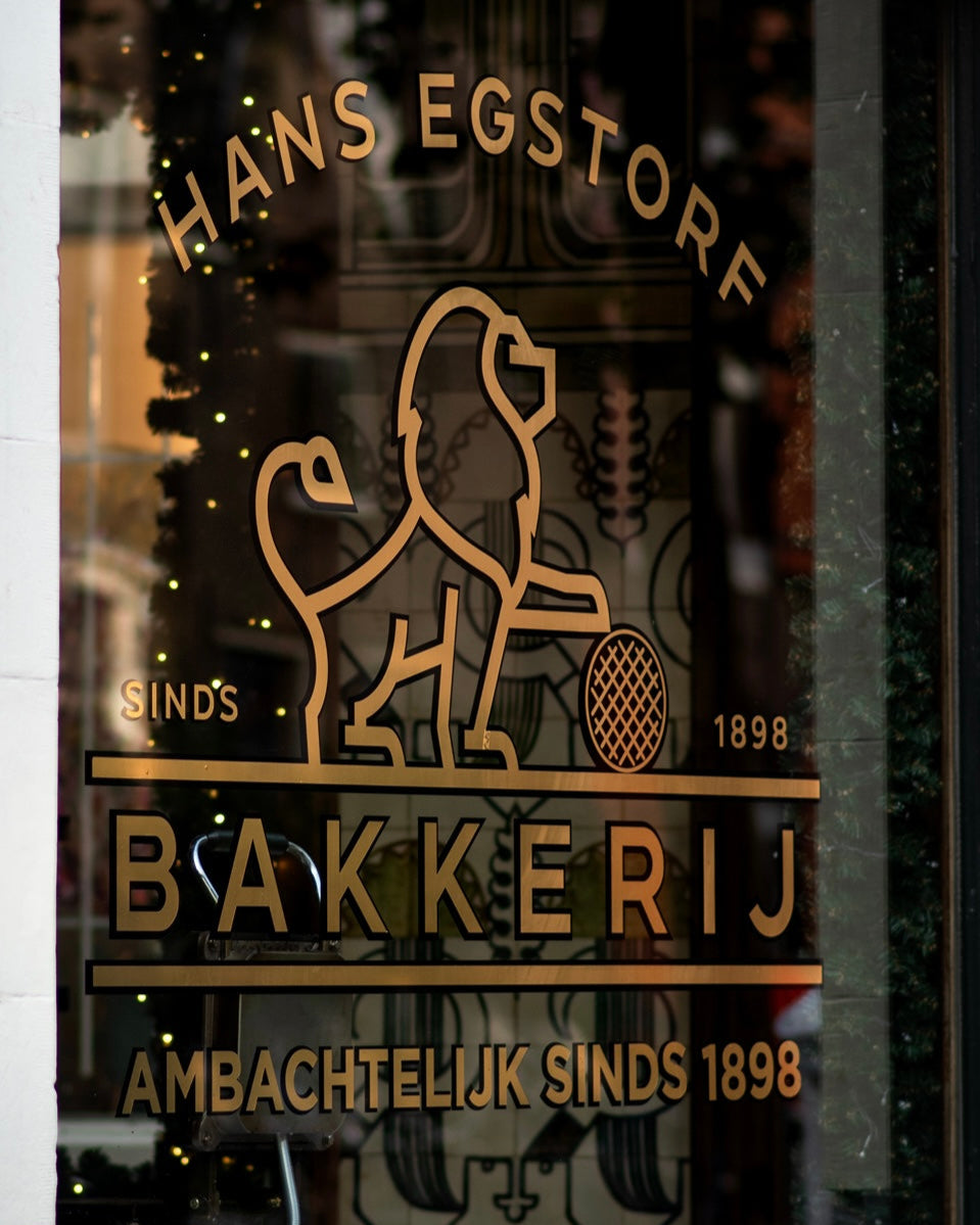 Glass door with gold logo and text of a bakery named 'Hans Egstorf Bakkery'.