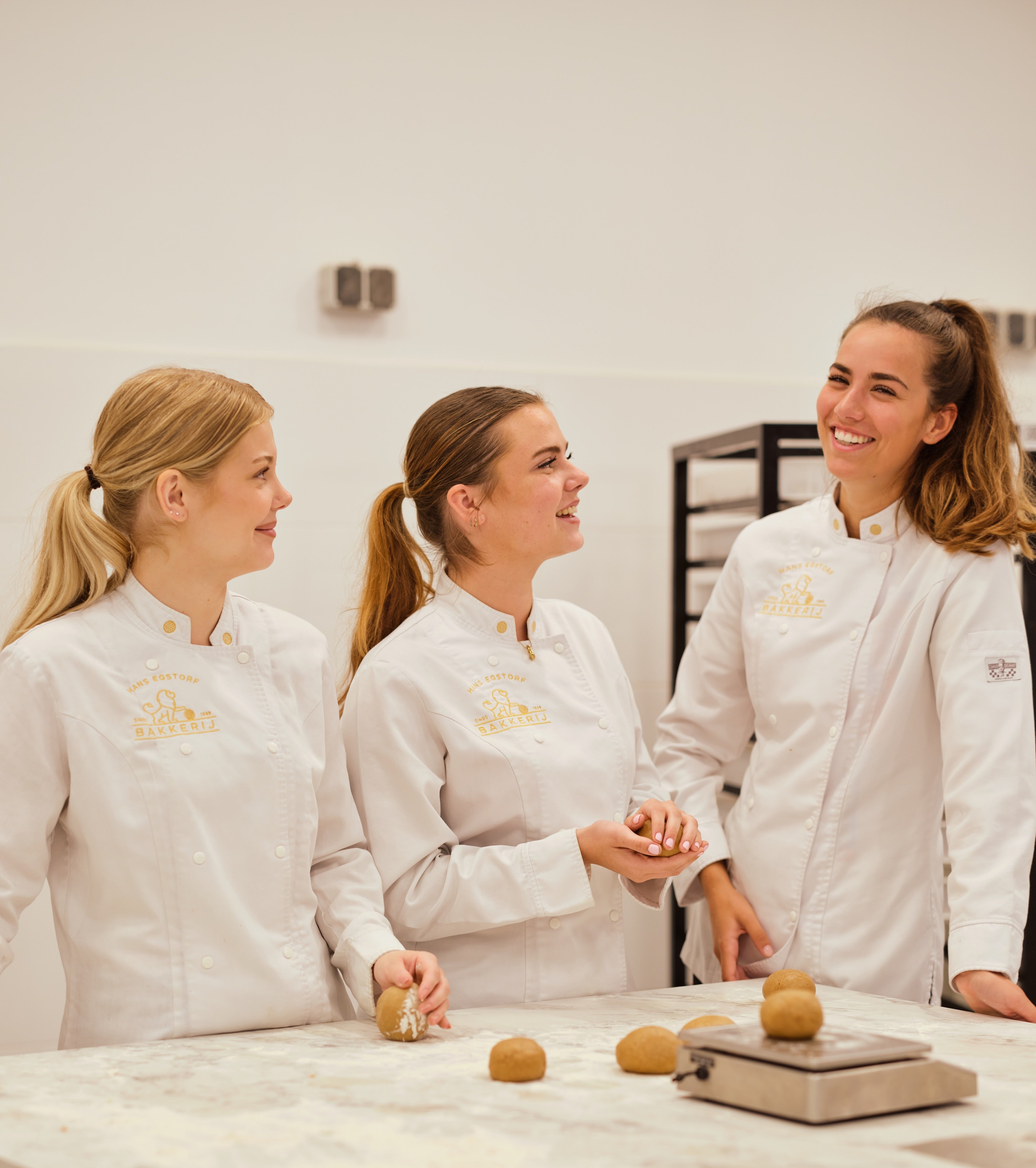 Three Hans Egstorf bakers, dressed in white chef coats, are gathered around a table crafting delicious Stroopwafels.